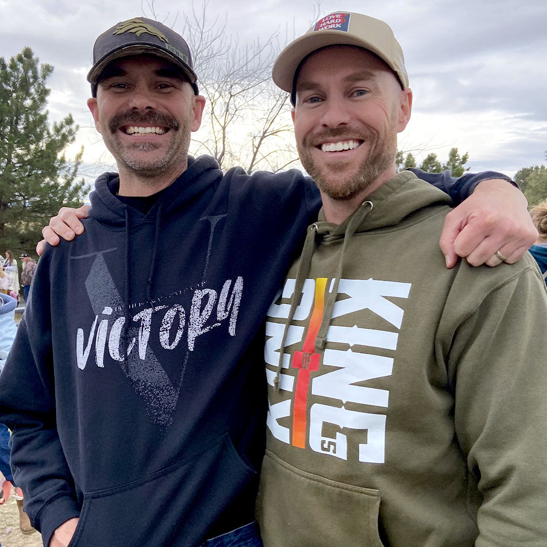 Two men wearing hoodies with 'Victory' and 'King of Kings' text, standing outdoors with trees and people in the background.